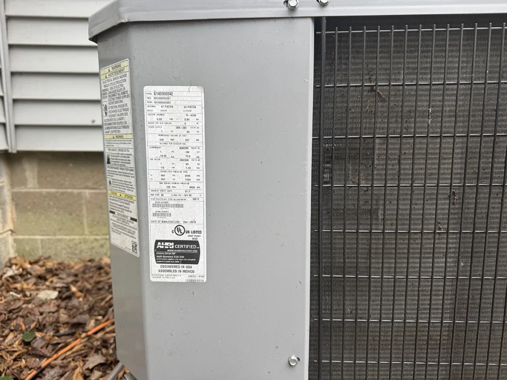 Close-up of the data label and condenser coil on an outdoor heat pump unit. Best for blogs about how to read HVAC model numbers, how to find tonnage on an AC unit, how old is my AC unit, HVAC serial number lookup, identifying your outdoor unit, and AC replacement planning.