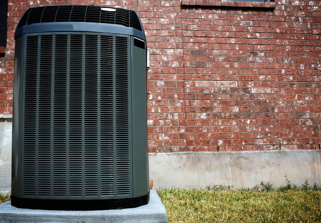 Outdoor residential heat pump unit installed on a concrete pad beside a brick home exterior. Best for blogs about heat pump systems, residential HVAC replacement, outdoor HVAC units, high-efficiency heating and cooling, heat pump installation, and AC vs heat pump topics.