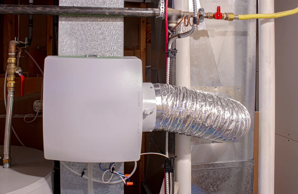 Whole-house humidifier mounted to ductwork with flexible bypass duct connected to a forced-air HVAC system. Best for blogs about whole-house humidifiers, dry indoor air, winter humidity control, and HVAC humidifier installation or maintenance.
